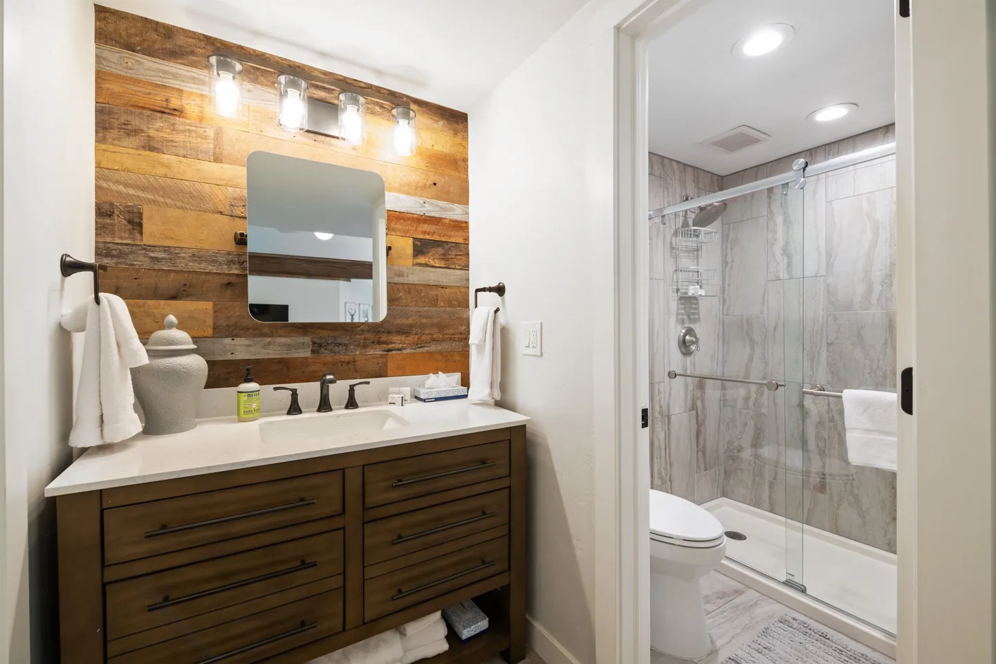 Bathroom vanity with wood-plank accent wall beside an open door leading to a toilet and glass-enclosed shower.