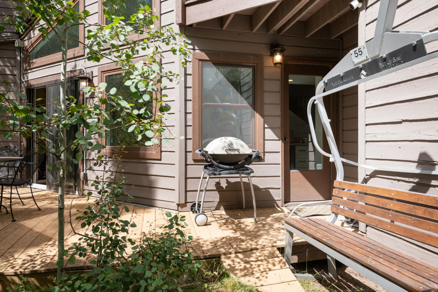 A wooden deck with a freestanding barbecue grill and a repurposed ski lift chair bench outside a home with wood siding.
