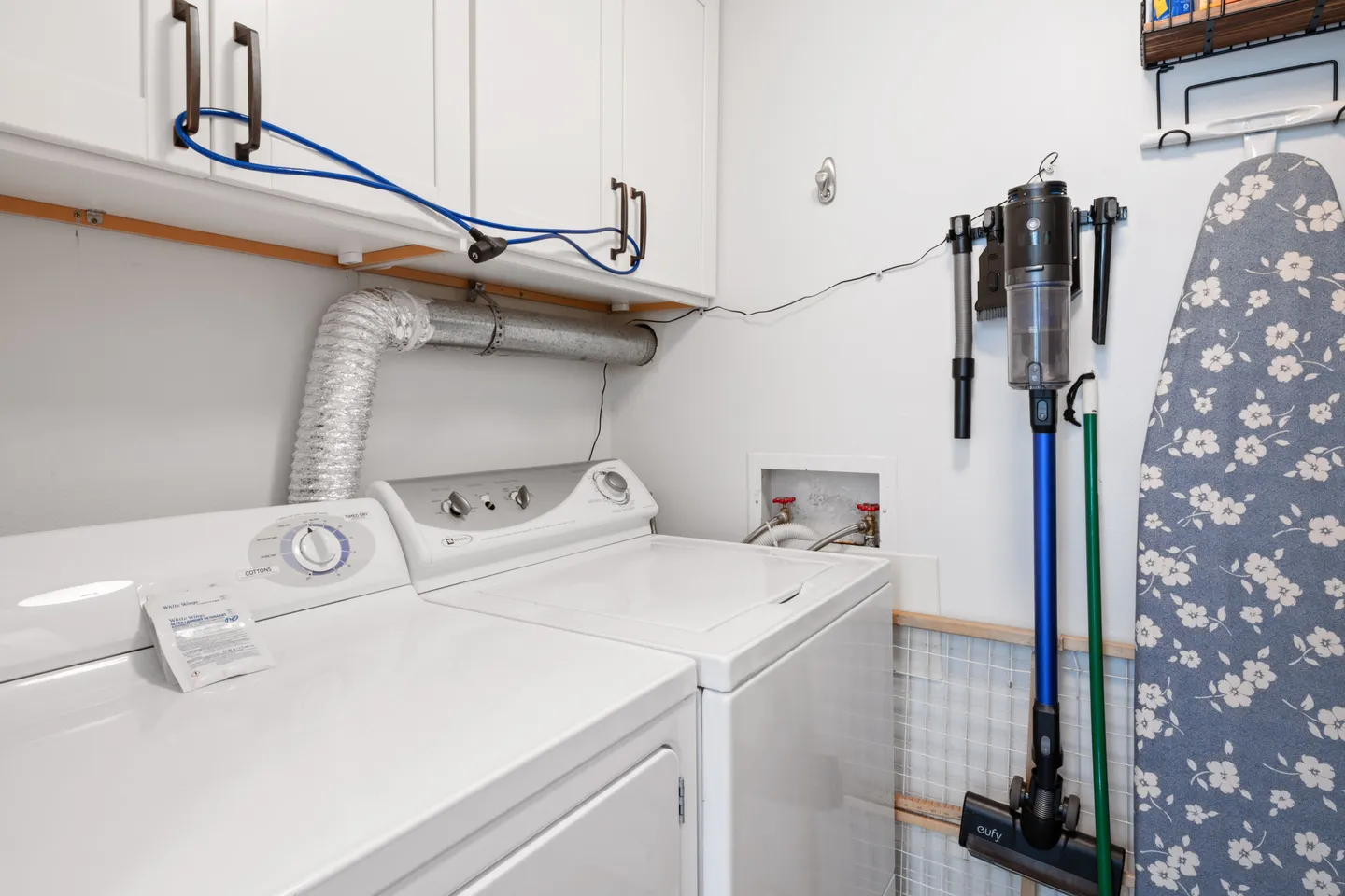 Laundry room with a white washer and dryer, upper cabinets, a wall-mounted vacuum, and an ironing board.