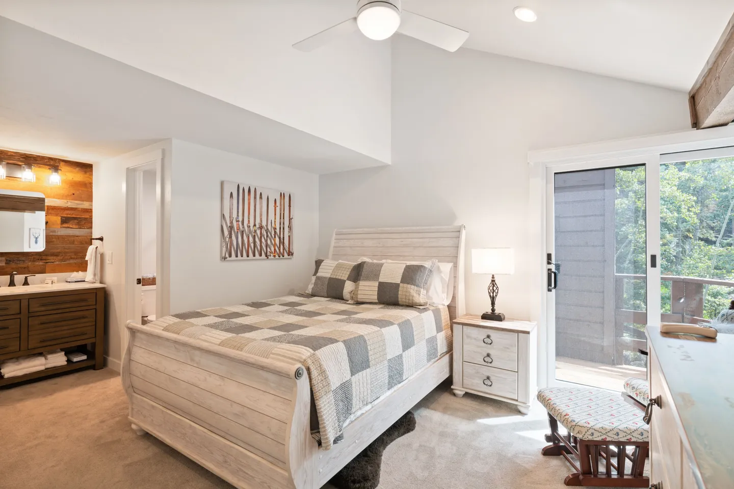 Bedroom with vaulted ceiling, light wood sleigh bed, ceiling fan, sliding glass door to a deck, and an ensuite vanity area.