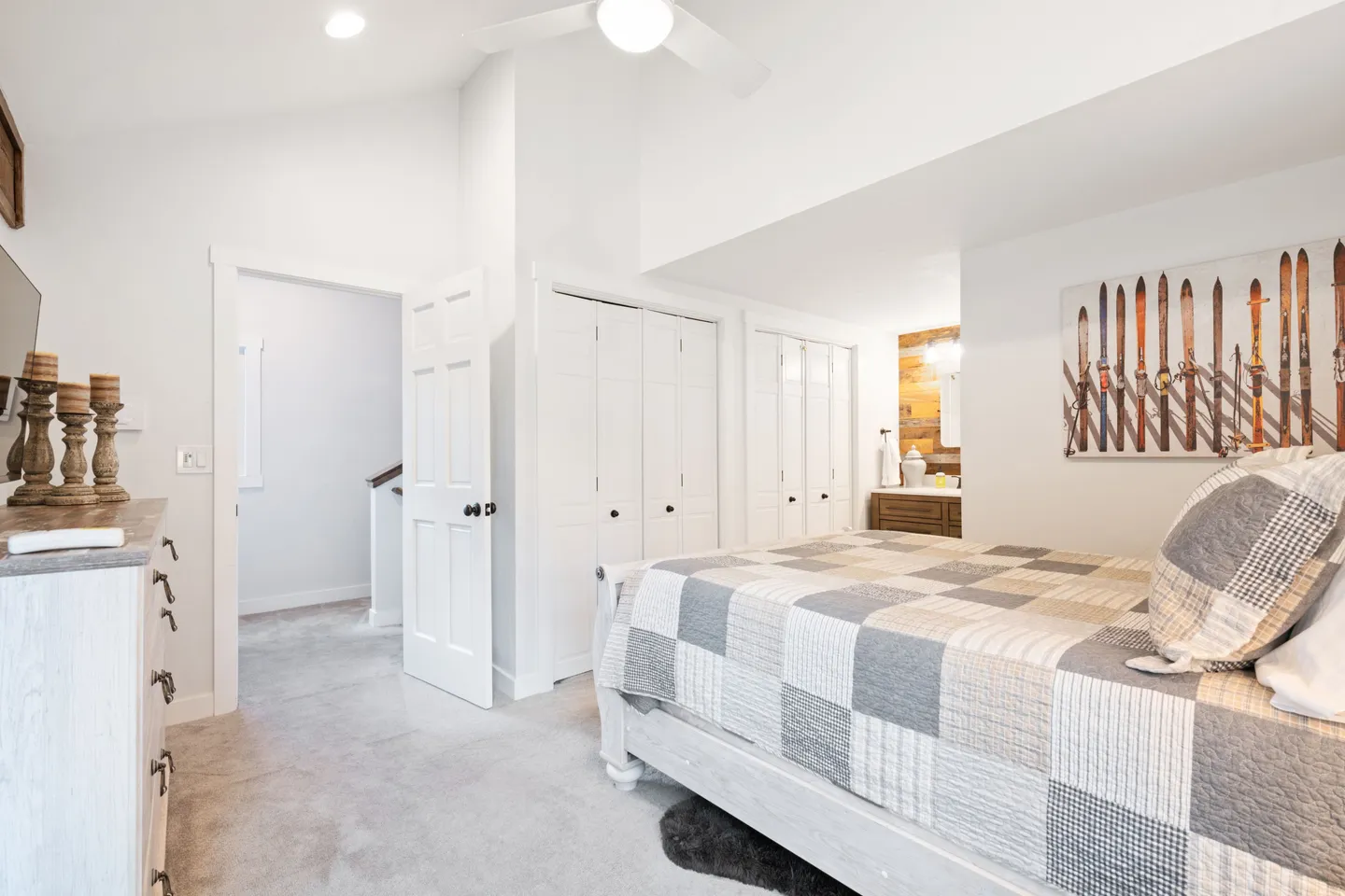 Bedroom with vaulted ceiling, light wooden bed with patterned quilt, ski-themed wall art, and an open doorway showing a glimpse of a bathroom.