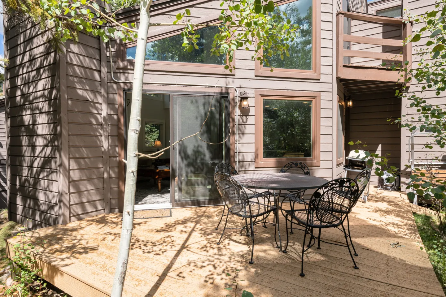 Sunlit wooden deck outside a brown wood-sided house, featuring a black wrought-iron patio table with chairs.