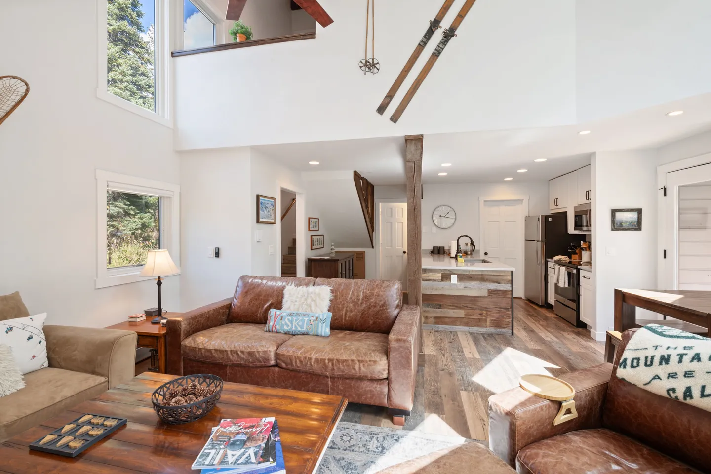 Open-concept living room with leather seating, wood coffee table, ski decor on the wall, and kitchen visible in the background.