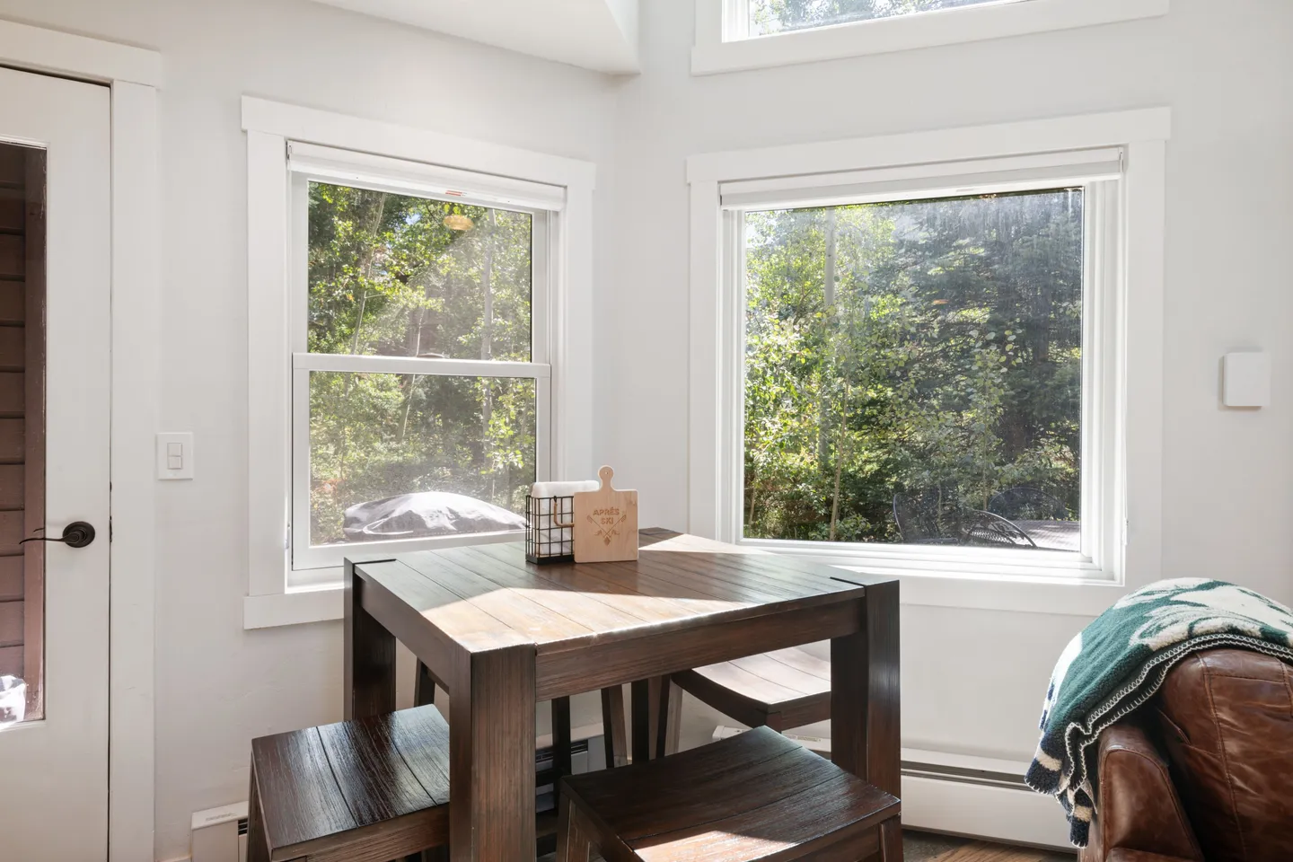 Dining nook with a dark wood table, benches, and stools surrounded by windows showing lush green trees.