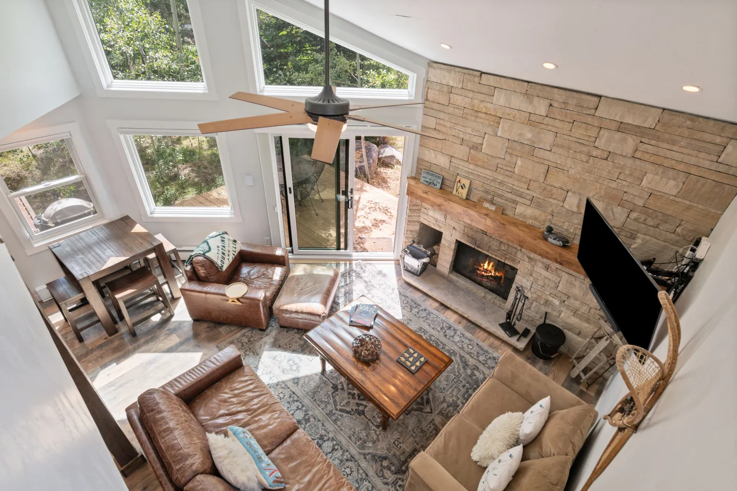High-angle view of a living room with a stone fireplace, leather and fabric seating, and tall windows.