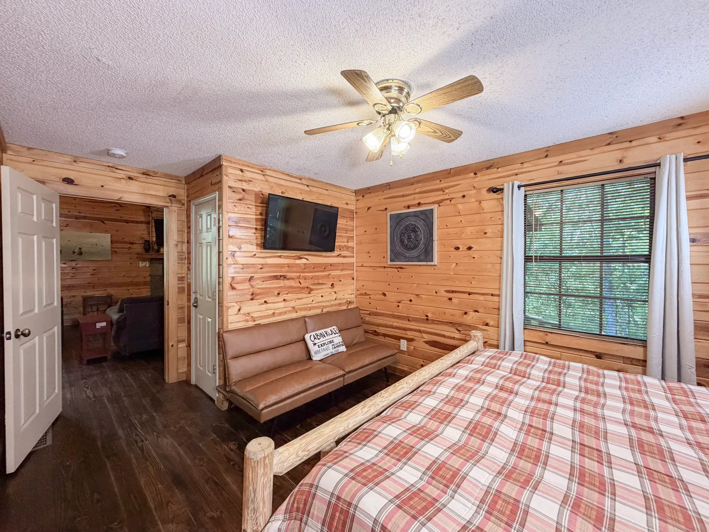 Bedroom with log walls, dark floors, a plaid-covered bed, a brown fold-out bench, a ceiling fan, and a wall-mounted TV.