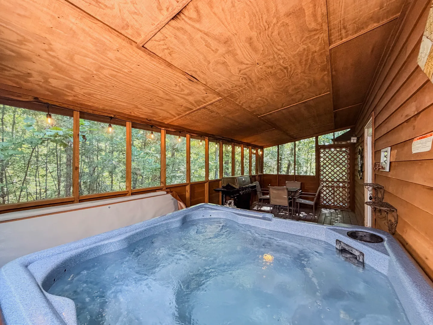 Screened-in porch featuring a bubbling hot tub, a dining set, and a barbecue grill with views of surrounding trees.