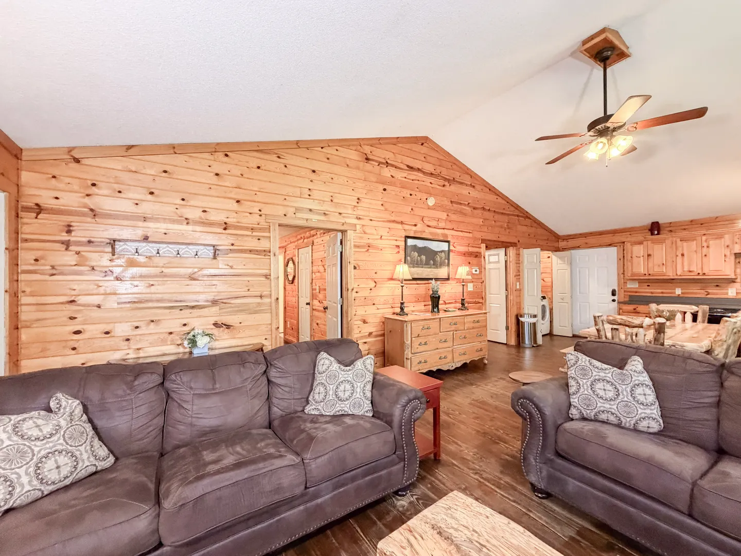 Open living area with wood-paneled walls, vaulted ceiling, brown plush sofas, and a view into the dining area and hallway.