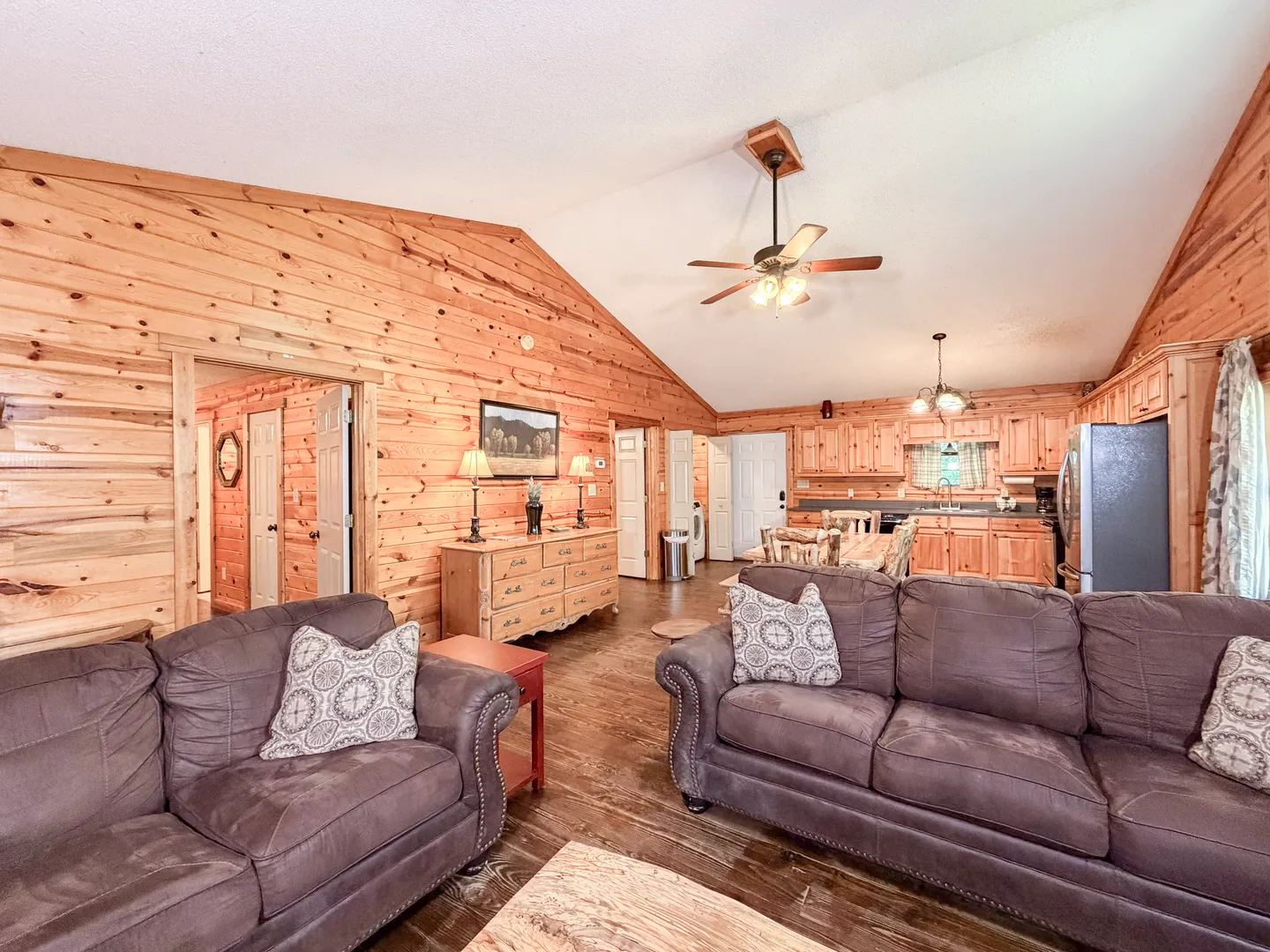 Open concept living room with plush couches, wood-paneled walls, vaulted ceiling, and view into the kitchen and dining area.