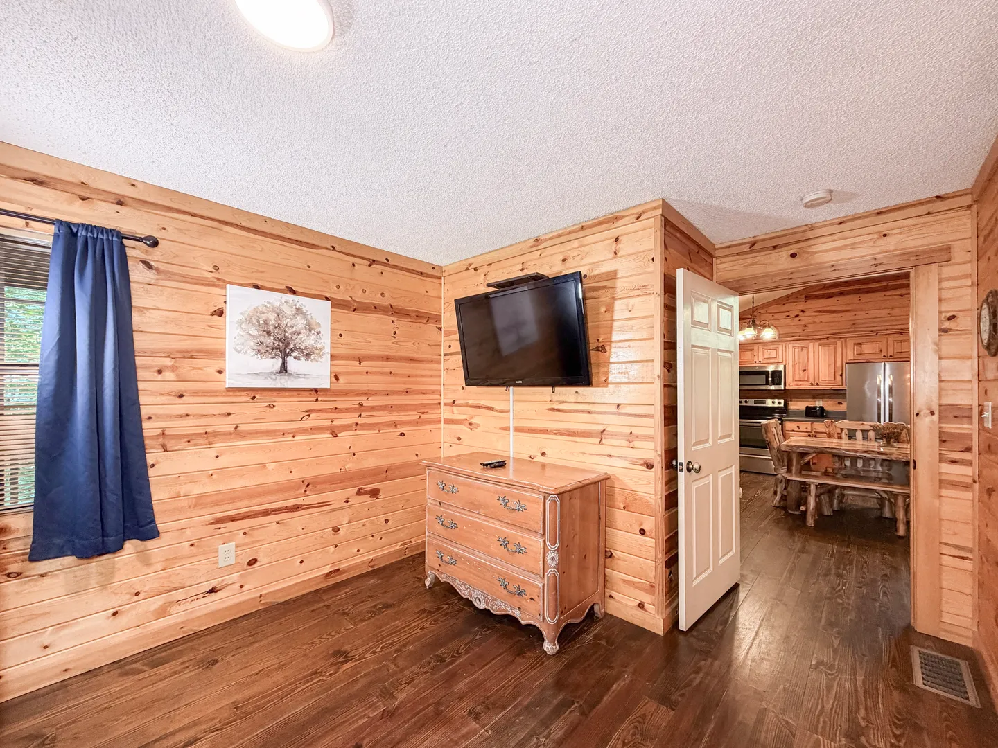 Bedroom with knotty pine wood paneled walls, a wall-mounted flat-screen TV above an ornate wooden dresser, and an open doorway leading to the kitchen and dining area.