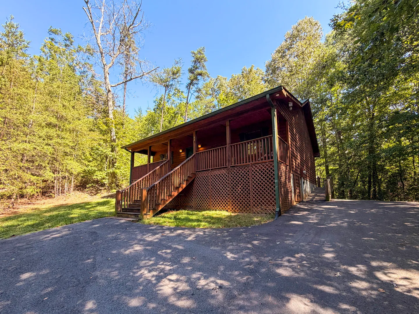 Exterior view of a wood cabin with a covered front porch, lattice skirting, and an asphalt driveway surrounded by lush green trees.