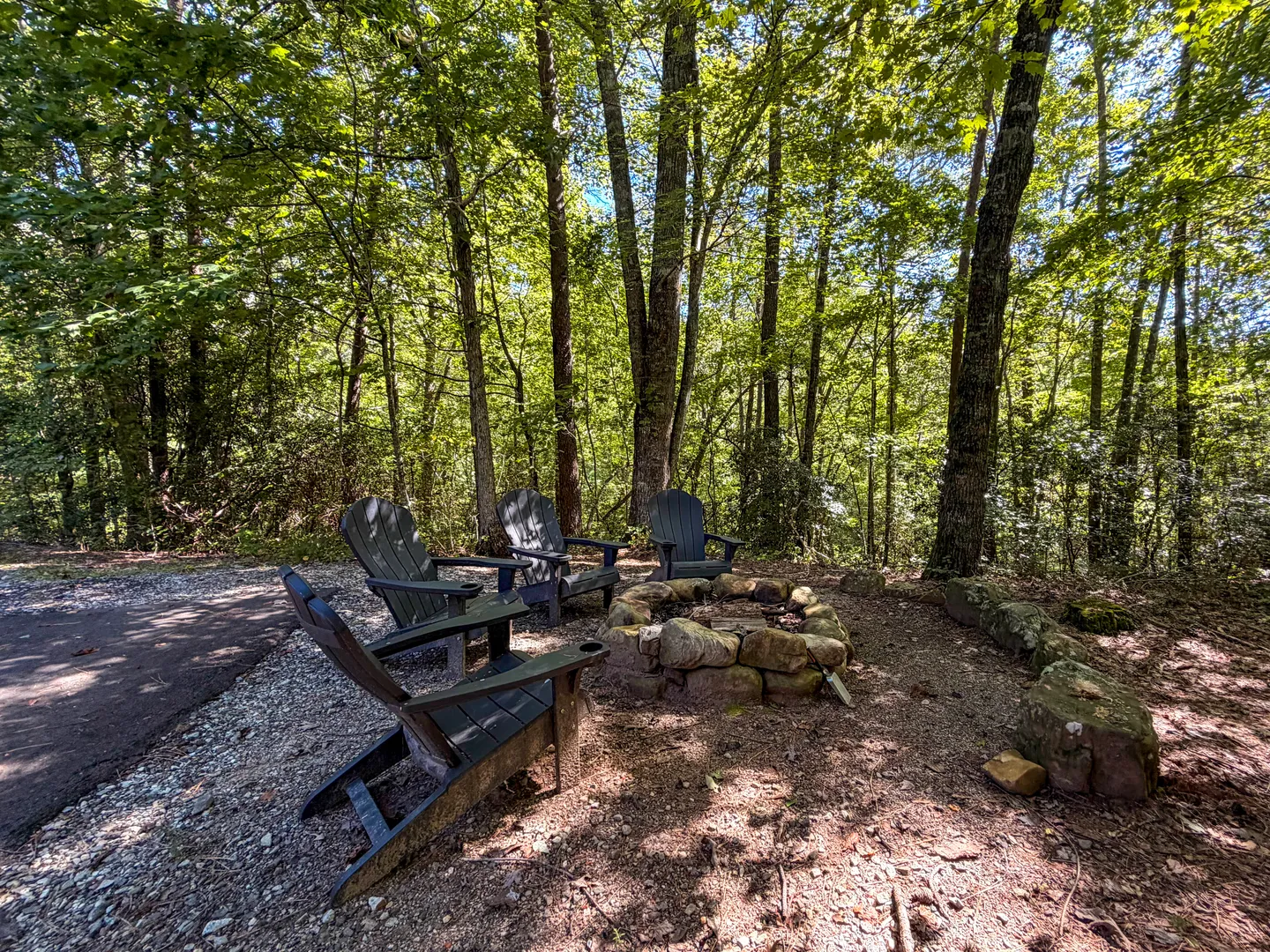 Outdoor fire pit surrounded by dark Adirondack chairs in a wooded setting next to a paved driveway.