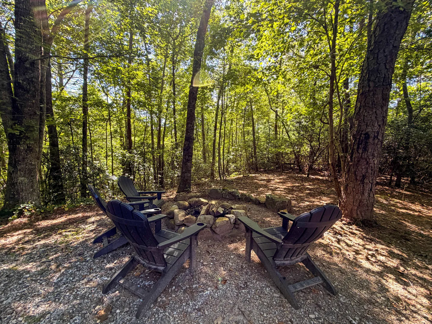 Outdoor fire pit surrounded by rocks and dark Adirondack chairs in a wooded setting.