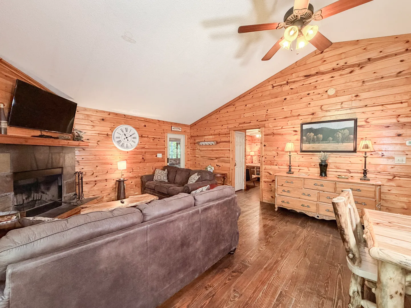 Living room with wood-paneled walls, a stone fireplace, brown sofas, and a vaulted ceiling with a fan.