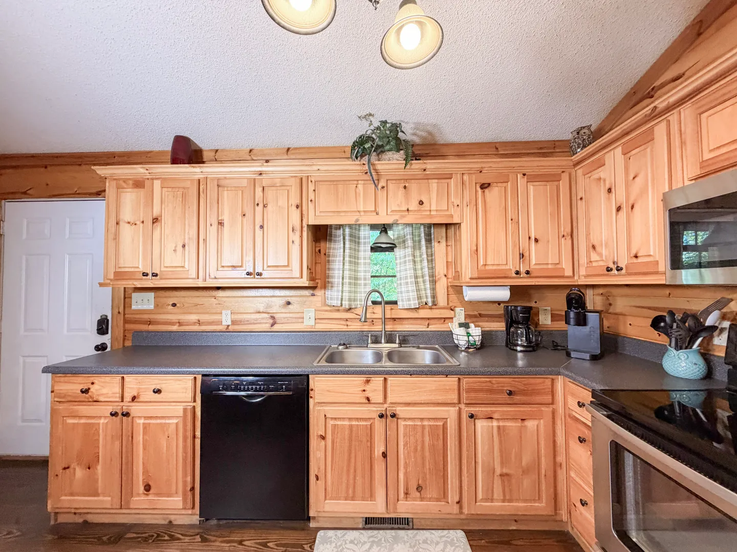 Kitchen with knotty pine cabinets, wood-paneled walls, dark countertops, a double sink, black dishwasher, and stainless steel oven.