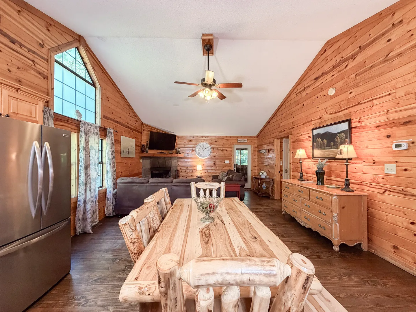 Open-concept cabin dining area with a rustic wood table leading into a living room with paneled wood walls and a vaulted ceiling.