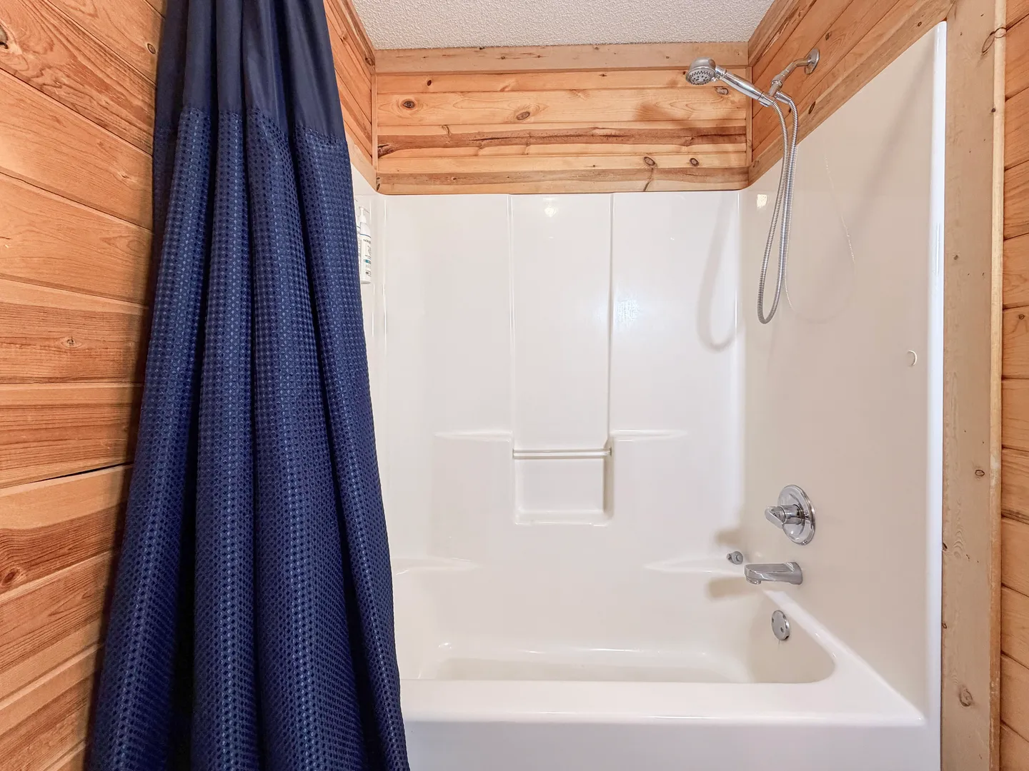 A bathtub and shower combination unit framed by wood-paneled walls with a dark blue waffle-weave shower curtain pushed to the side.
