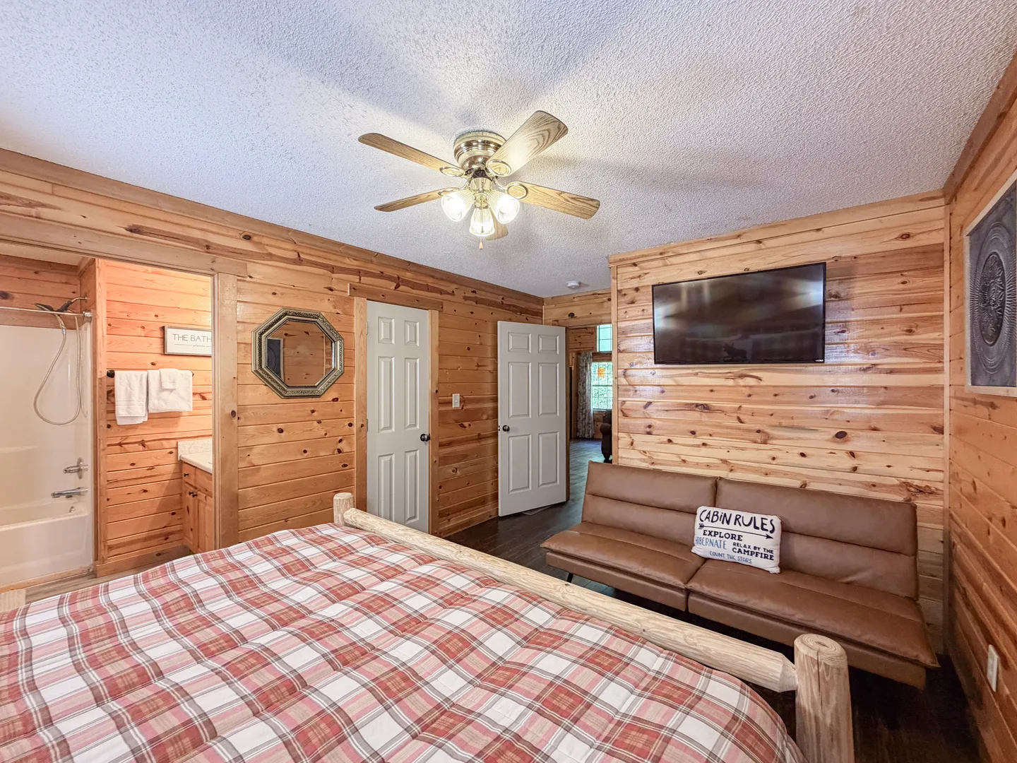 Bedroom with log bed frame, plaid bedding, wood-paneled walls, ceiling fan, brown futon sofa, wall-mounted TV, and an adjoining bathroom.