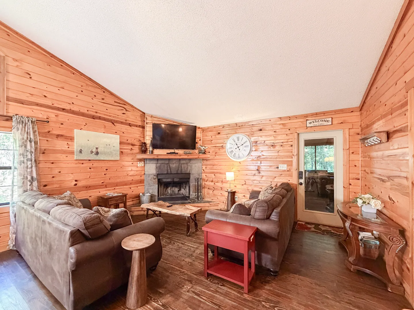 Living room with knotty wood-paneled walls, wood floors, a corner stone fireplace with a mounted television, and plush sofas.