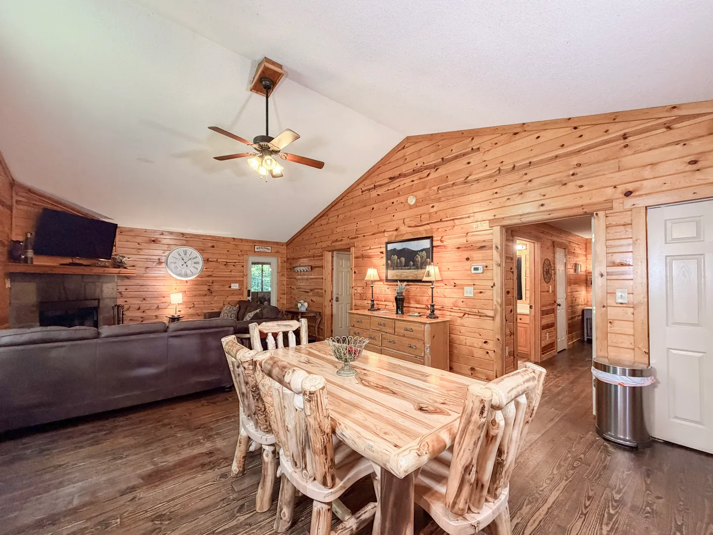 Open-concept cabin interior showing a log-style wooden dining set in the foreground and a living room with a stone fireplace in the background.
