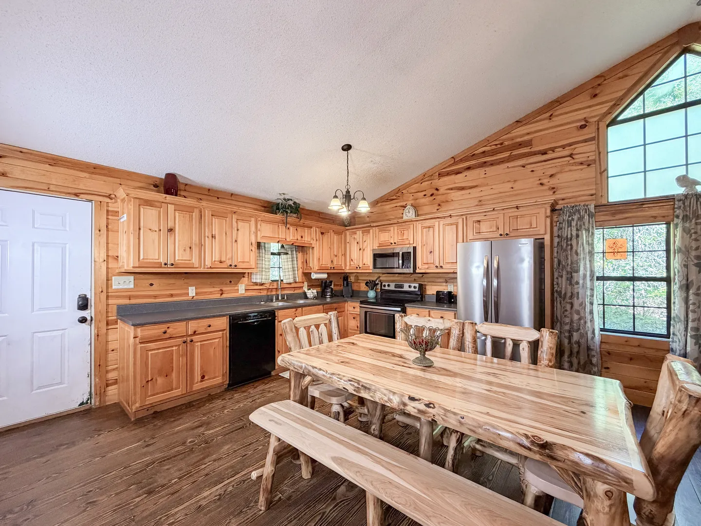 Open dining and kitchen area with natural wood cabinetry, a rustic log dining table, and wood-paneled walls under a vaulted ceiling.