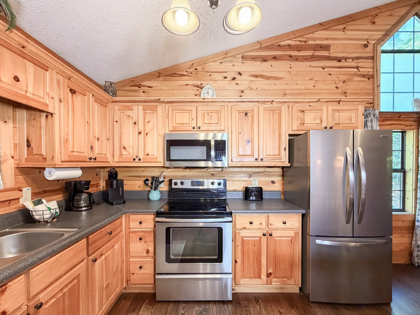 Kitchen with natural wood cabinets, stainless steel appliances, dark countertops, and a vaulted wood-plank wall.