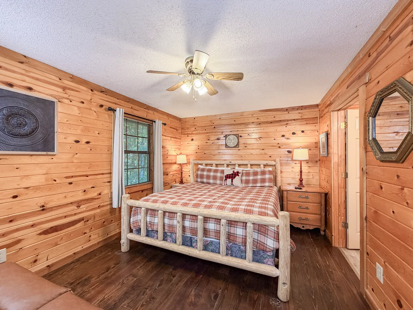 Bedroom with log-style bed frame, knotty wood-paneled walls, ceiling fan, and dark flooring.