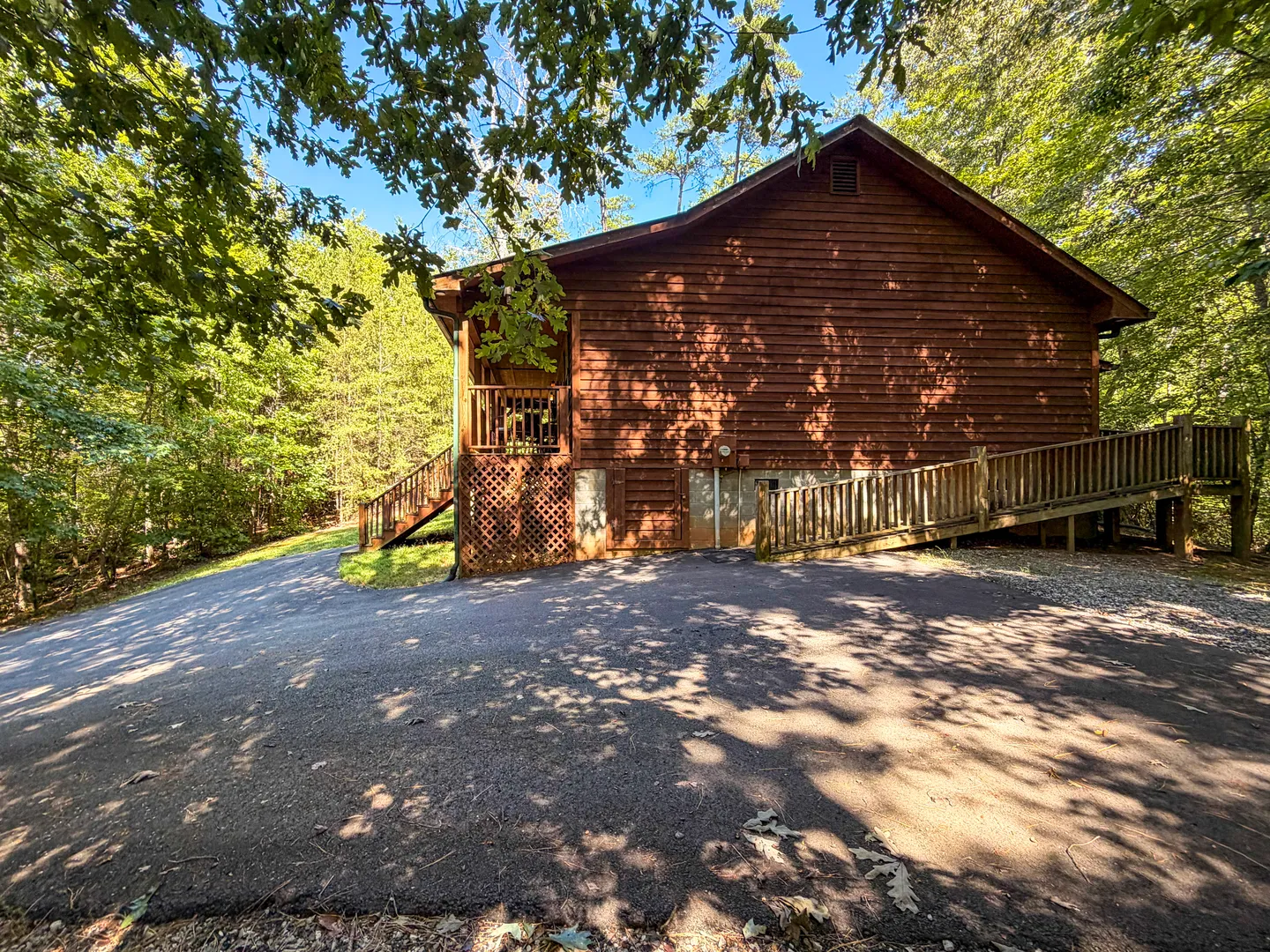 Exterior side view of a brown wooden log-sided cabin with a paved driveway and ramp entry surrounded by green trees.