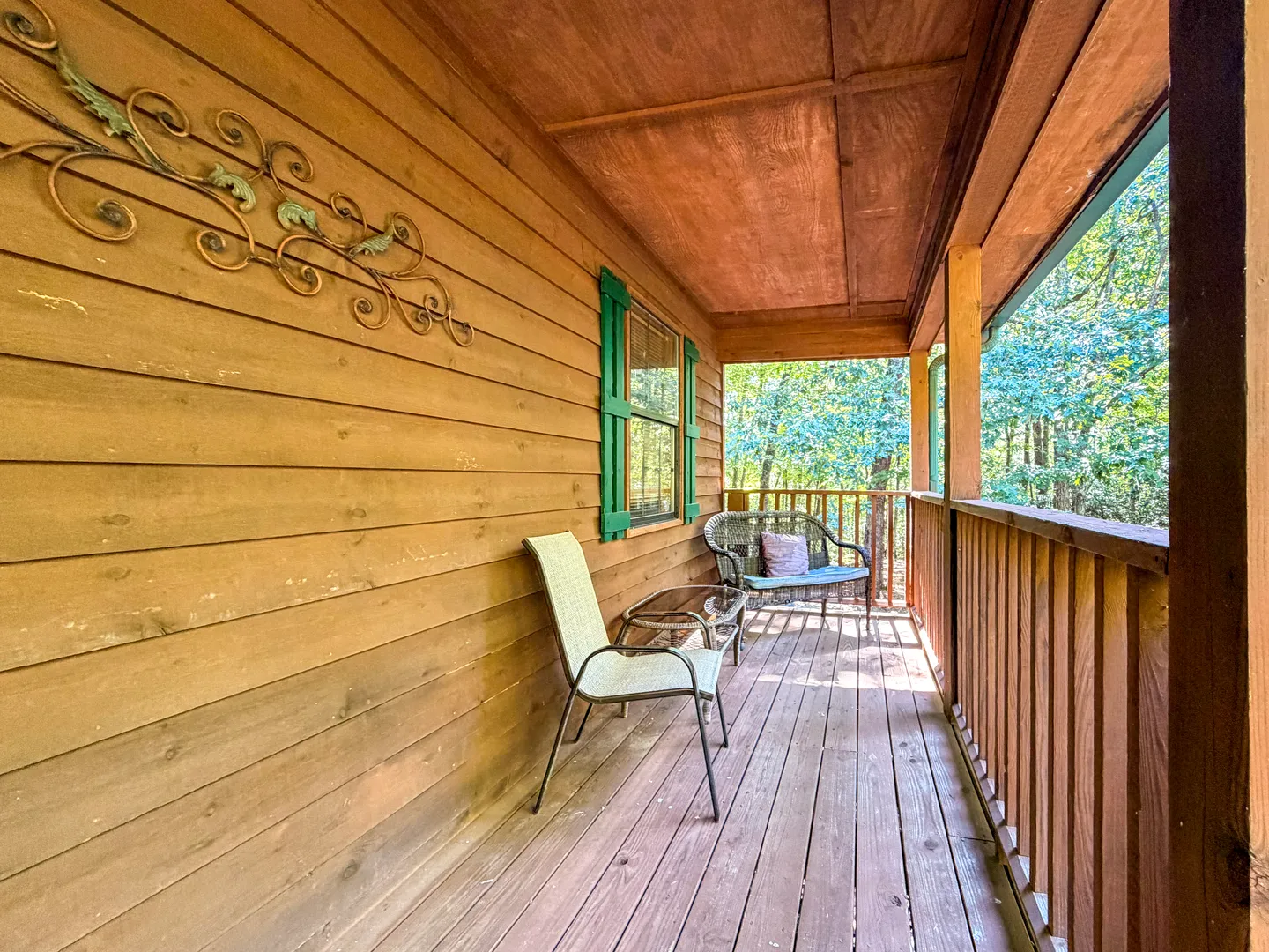 A covered wooden porch along a log cabin exterior, featuring a chair, small table, and a loveseat bench with views of surrounding trees.