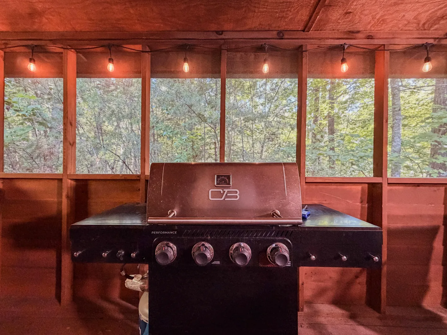 A barbecue grill situated on a screened-in porch with string lights hanging overhead and woods visible outside.