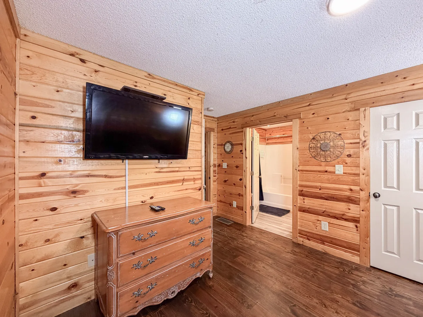 Bedroom with log siding walls, a mounted TV over a wooden dresser, and an open doorway leading to an adjoining bathroom.