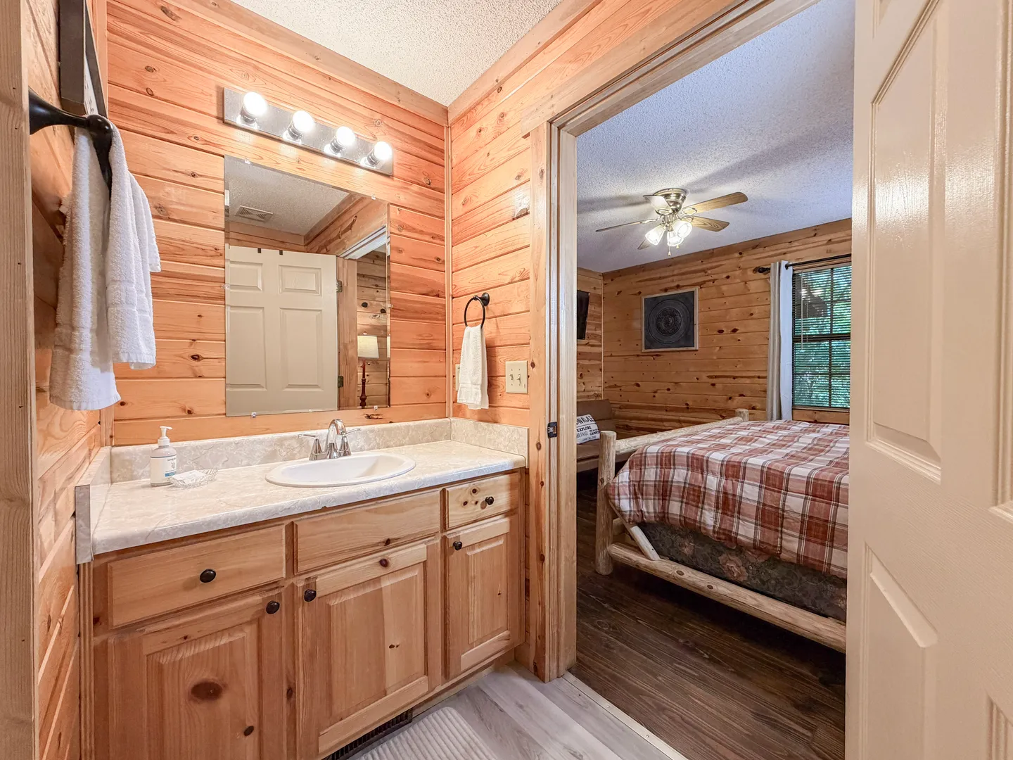 Bathroom with wood-paneled walls and a wooden vanity looking into an adjoining bedroom with log-style furniture.