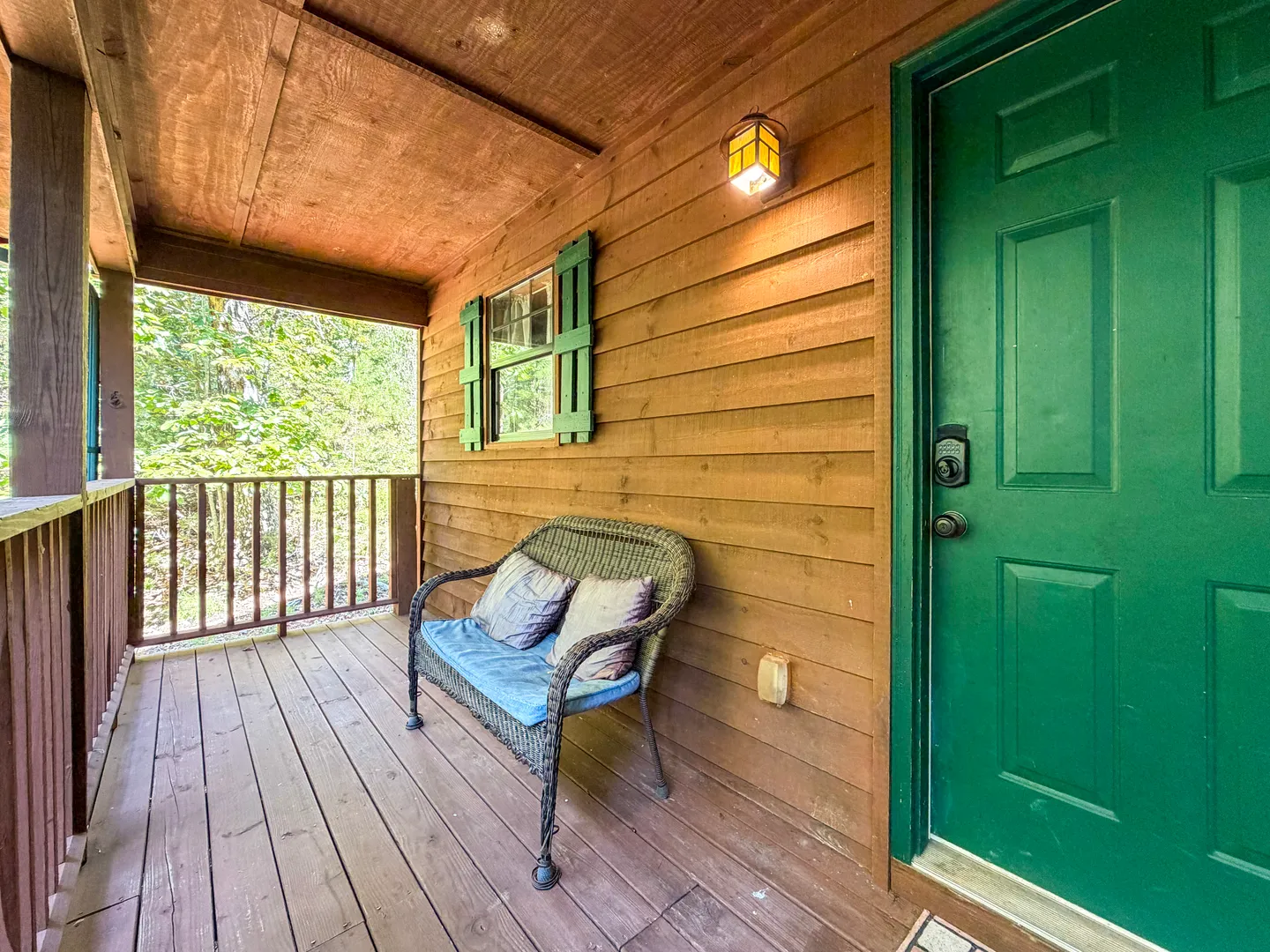 Covered wooden porch with a green front door, a wicker loveseat bench, and a small shuttered window.