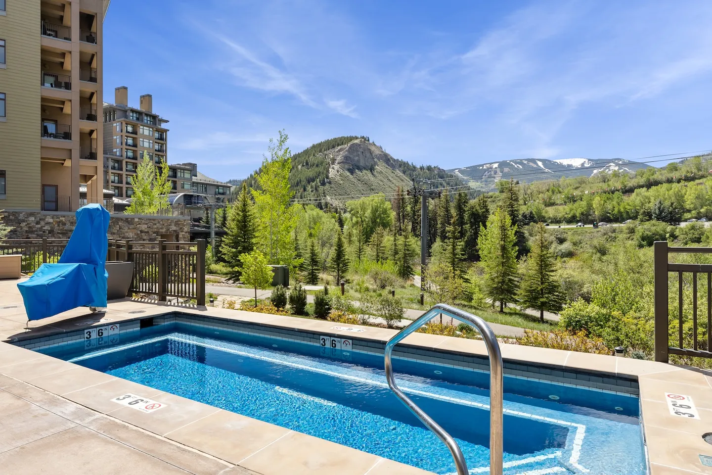 Outdoor rectangular hot tub or plunge pool on a patio overlooking mountains, evergreen trees, and neighboring lodge buildings under a clear blue sky.