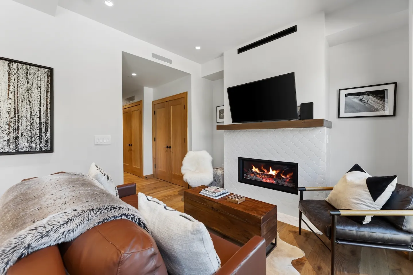 Living room with a modern fireplace, wall-mounted TV, leather couch, and armchairs on wood flooring.