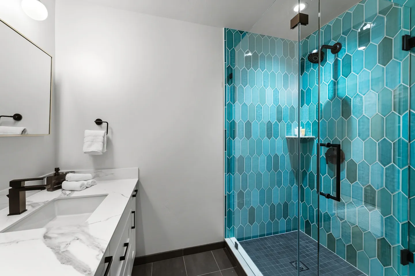 Bathroom vanity with marble countertop next to a walk-in shower with turquoise picket tile and a glass door.