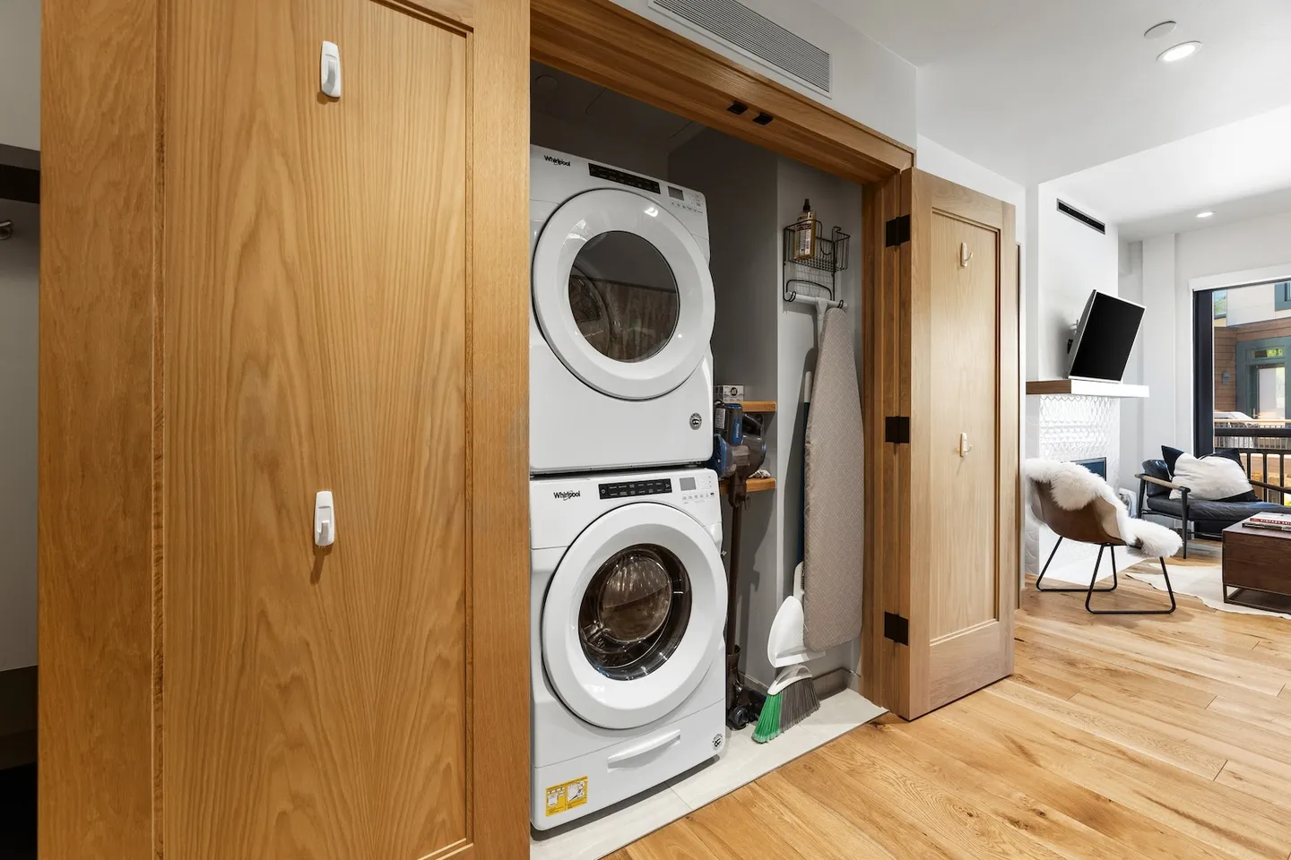 Open closet with stacked white washer and dryer next to cleaning supplies, with wood floors leading to an adjacent living room.