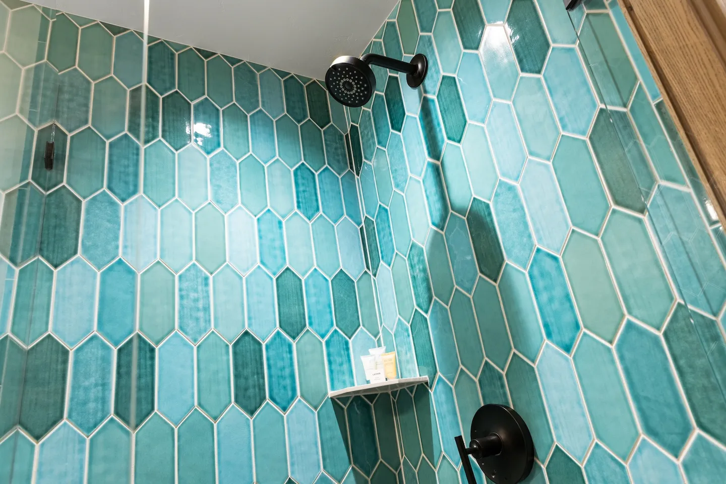 Walk-in shower featuring elongated hexagonal teal tiles, a black showerhead, and a corner shelf with toiletries.
