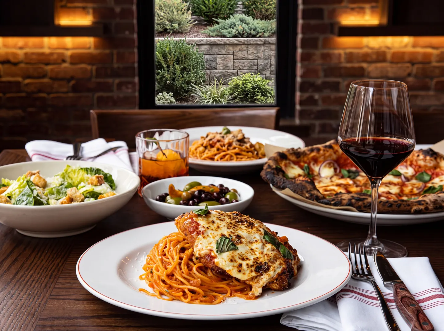 A wooden table set with an Italian meal including chicken parmigiana over pasta, pizza, salad, olives, and a glass of red wine in front of a brick wall and window.