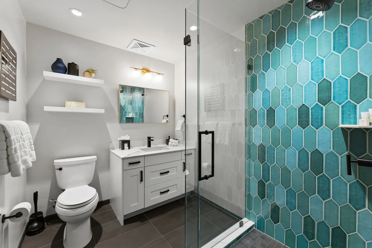 Bathroom featuring a glass-enclosed walk-in shower with turquoise elongated hexagon wall tiles, a white vanity with black hardware, and a toilet.