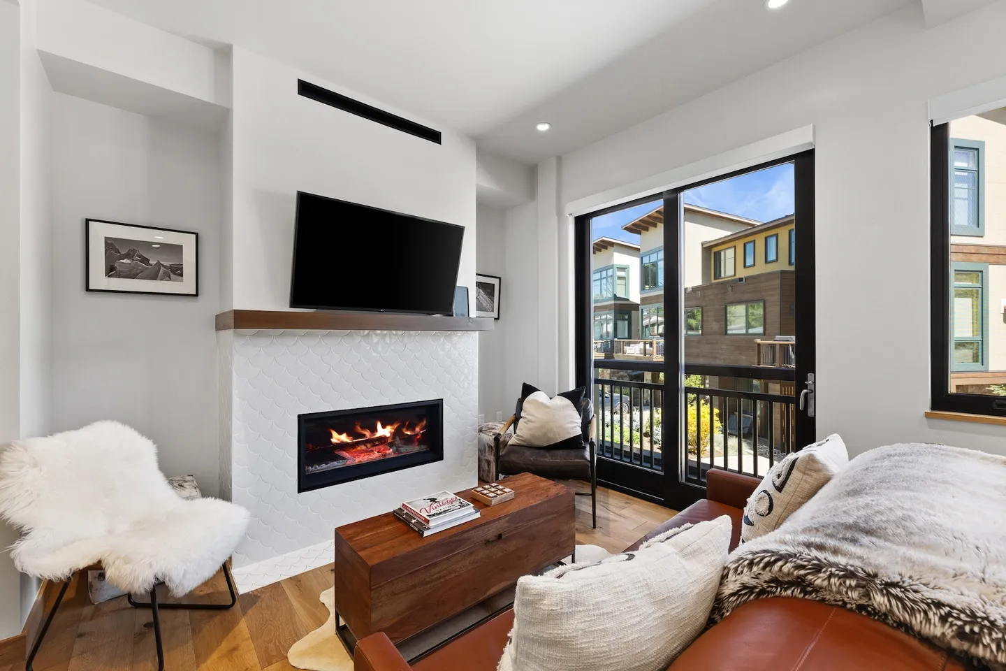 Living room with a tiled fireplace, mounted TV, leather sofa, rustic coffee table, and sliding glass doors to a balcony.