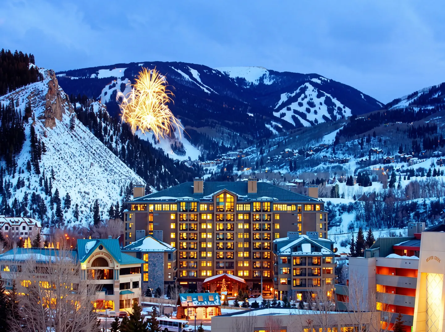 Dusk view of a multi-story mountain lodge illuminated with warm lights beneath snowy peaks and fireworks in the evening sky.