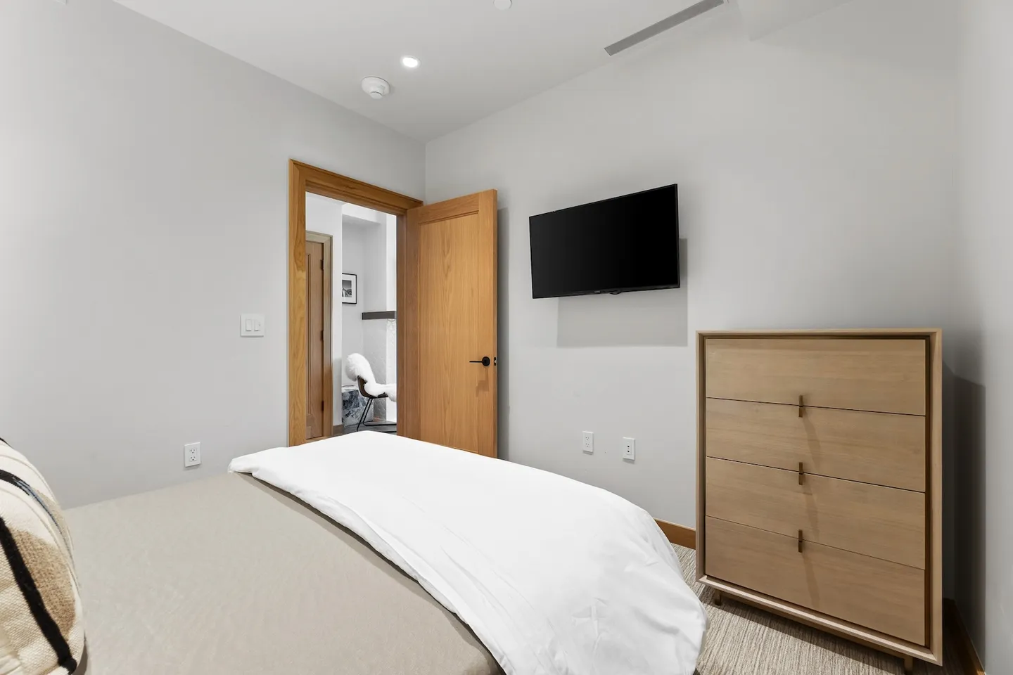 Bedroom with a bed in the foreground, a wall-mounted flat-screen TV, a light wood chest of drawers, and an open doorway showing an adjoining room.