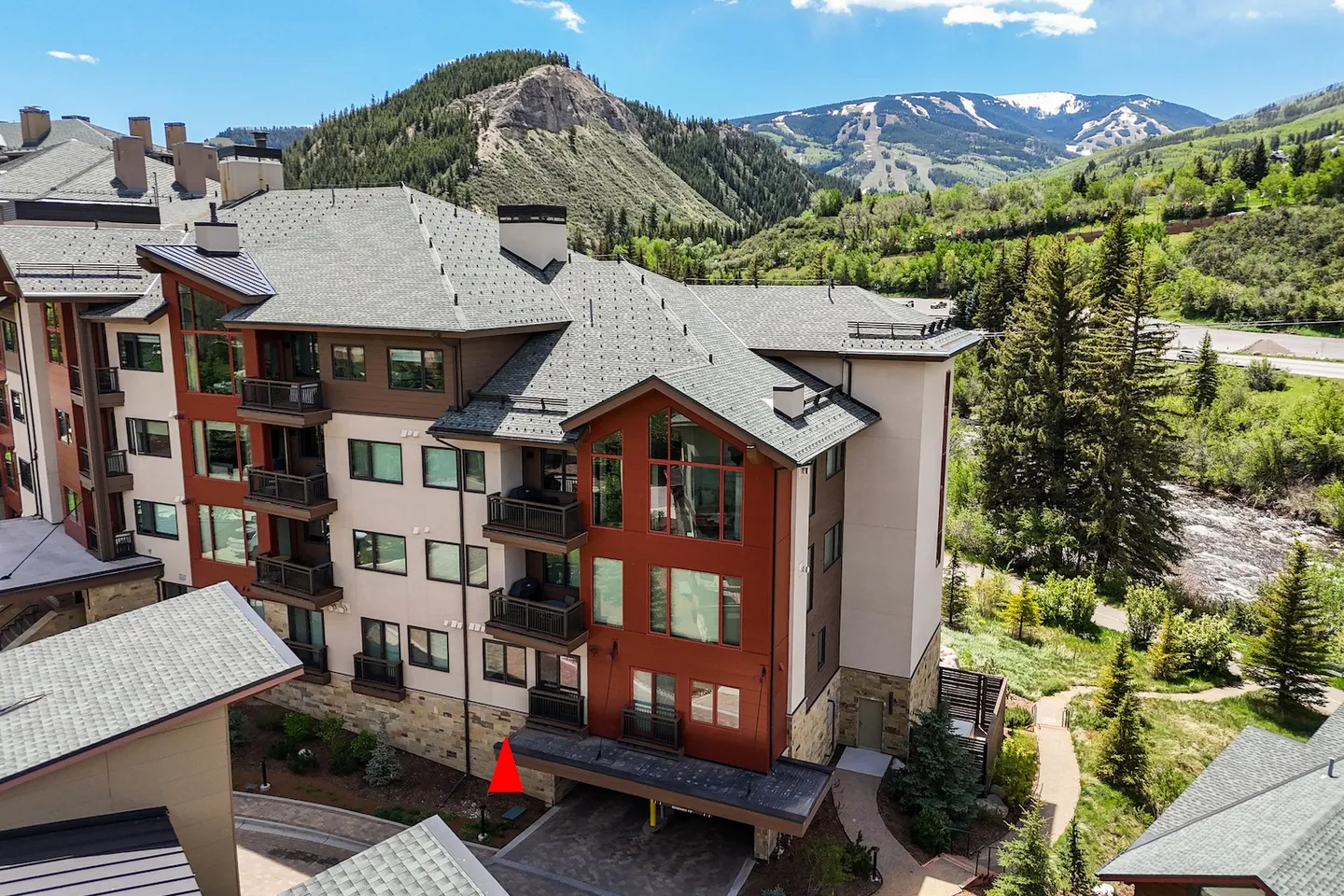 Aerial view of a multi-story lodge-style condominium building surrounded by pine trees with snow-capped mountains in the background under a blue sky.
