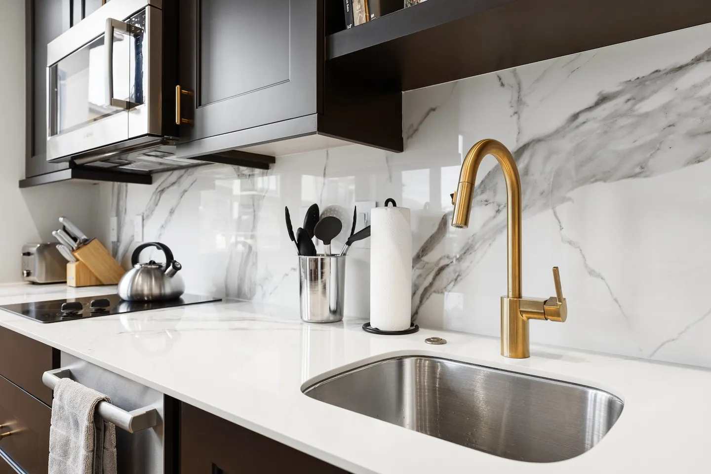 Close-up of a kitchen countertop with an undermount stainless steel sink, gold gooseneck faucet, and marble-patterned backsplash.