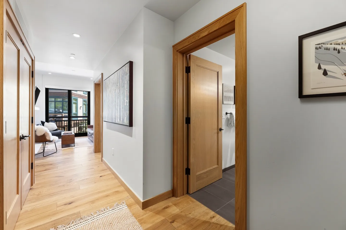 Hallway with wood floors, light-colored walls, wood doors and trim, leading toward a bright living room.