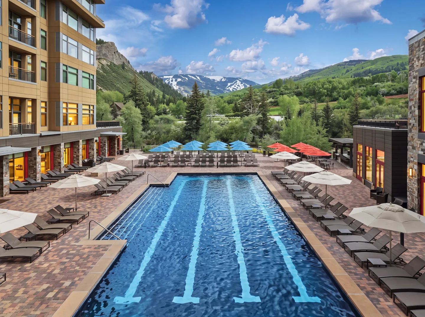 An outdoor lap swimming pool surrounded by paved decking, lounge chairs, and umbrellas with a mountain backdrop.
