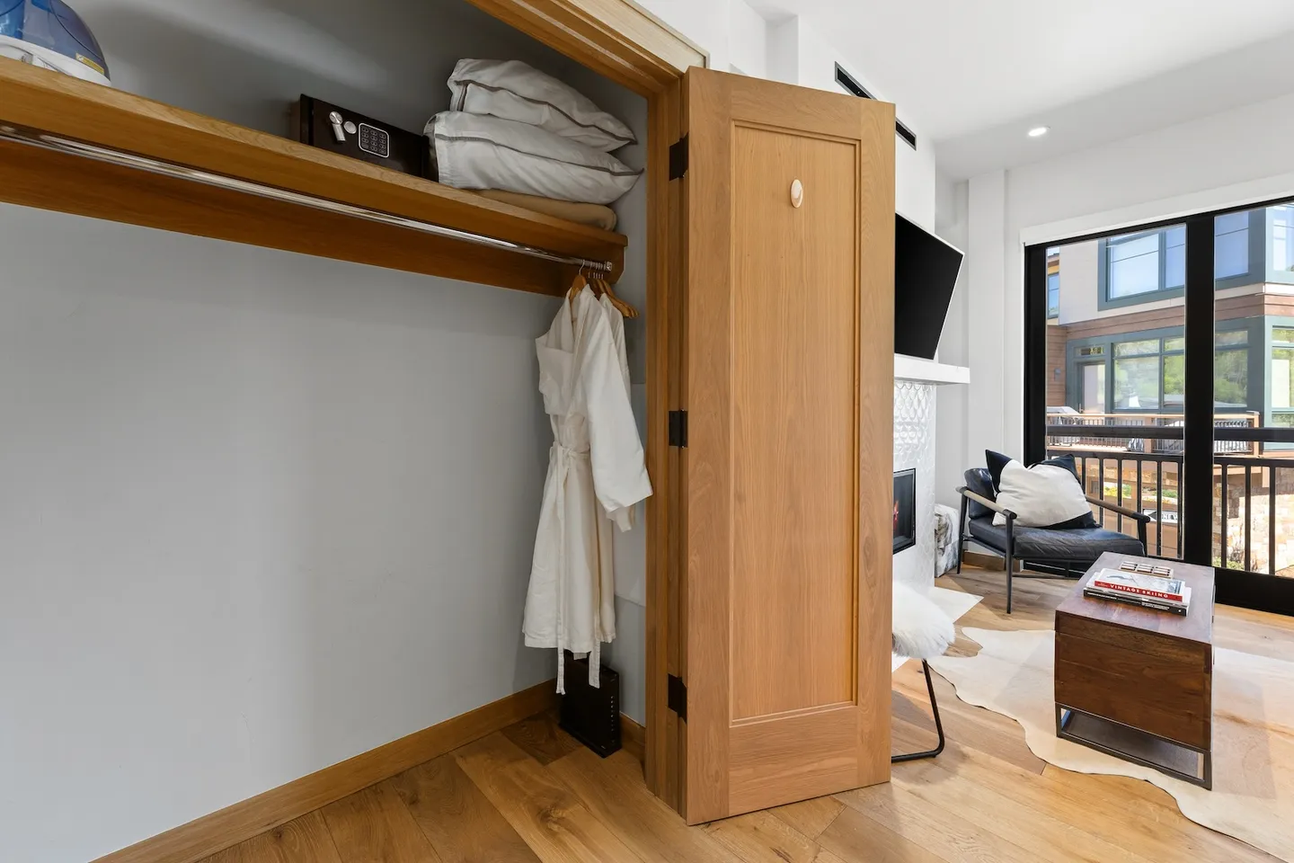 Open wooden closet with bathrobes, safe, and bedding, adjacent to a living area with a fireplace and balcony door.