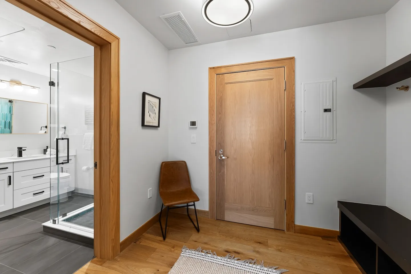 Entryway with light wood door, wood flooring, brown chair, and view into an adjacent bathroom with glass shower.