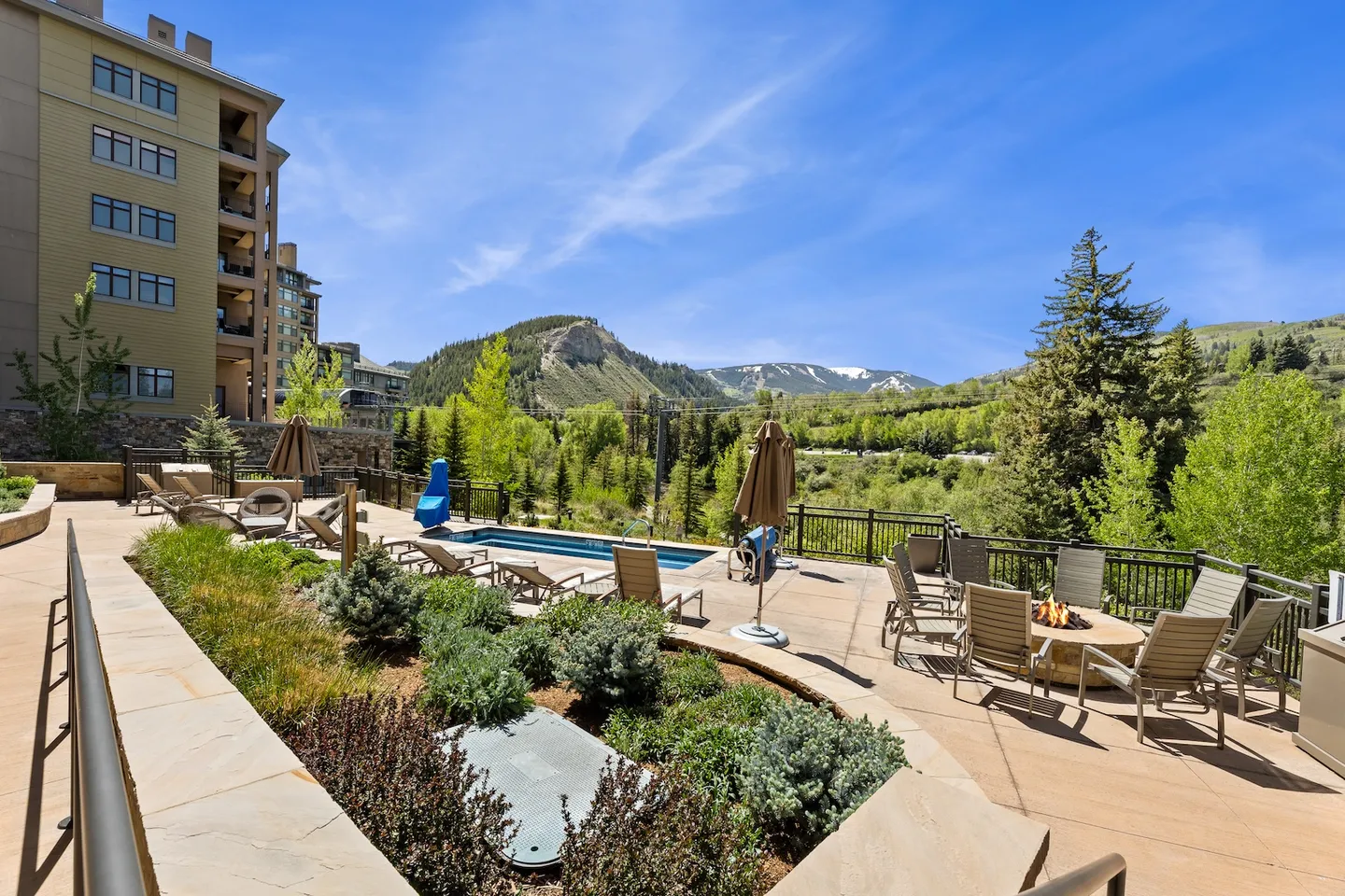 Outdoor patio with a swimming pool, lounge chairs, a fire pit area, and views of forested mountains under a blue sky.